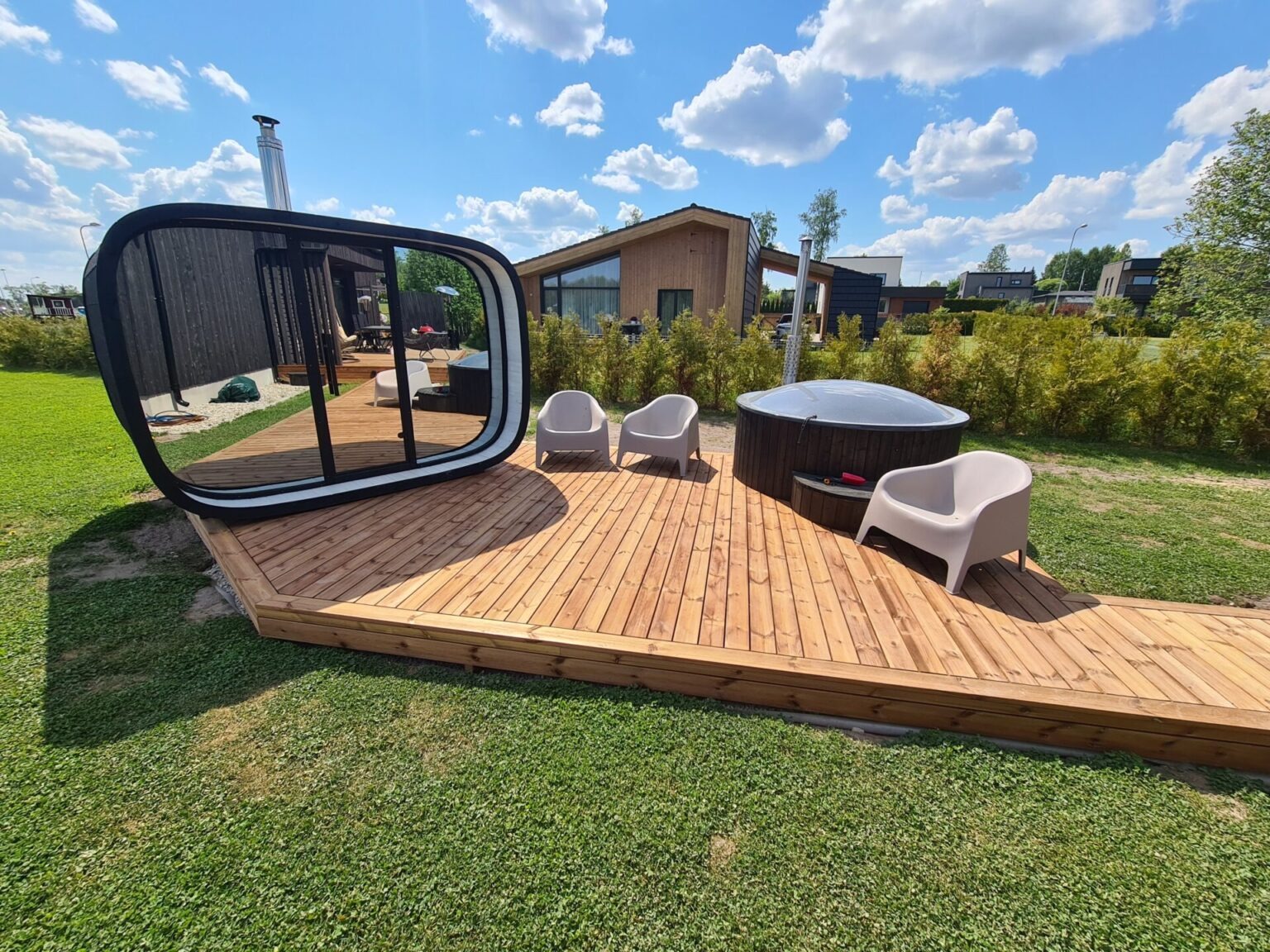 Kaasaegne välikompleks puidust terrassil – ovaalne peegelpinnaga saun, kümblustünn ja kaks valget tooli rohelisel murul päikeselise ilmaga.