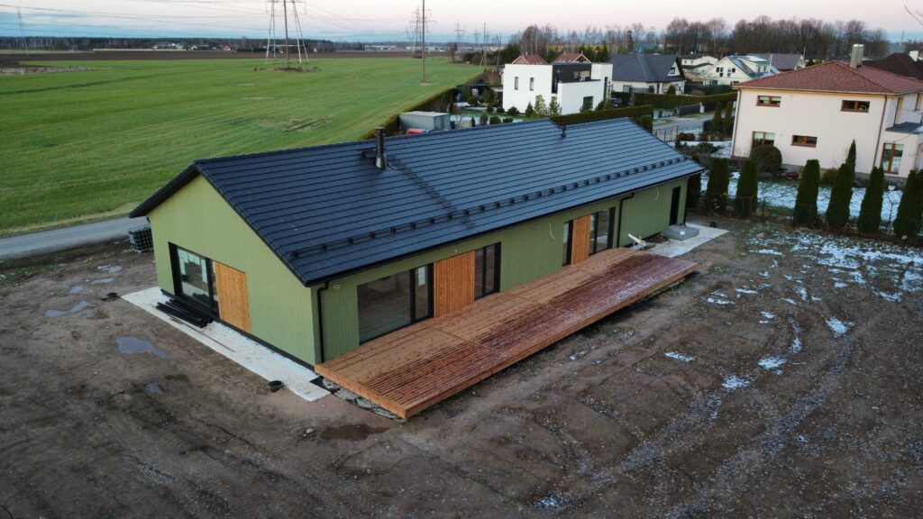 Roheline ühekorruseline puitfassaadiga maja musta kivikatuse ja suure puidust terrassiga, paiknedes avatud maastikul ja kaasaegses elamurajoonis.