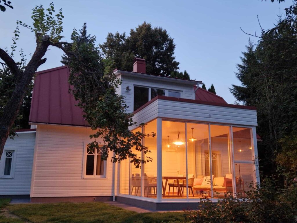 Valge puitmaja punase katuse ja valgusküllase klaasverandaga, mille soe sisevalgus loob õhtuses aias hubase meeleolu.