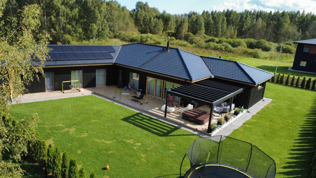 Kaasaegne ühekorruseline musta fassaadiga maja, millel on päikesepaneelid katusel, suur haljastatud aed, terrass pergolaga ja pere puhkeala koos batuudi ja aiamööbliga.