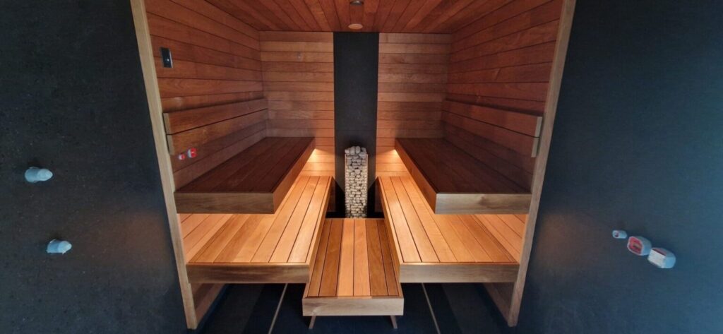 Kaasaegne puidust saun kahe vastastikuse astmelise lavaga ja keskel paikneva kivikerisega tumeda seina taustal.