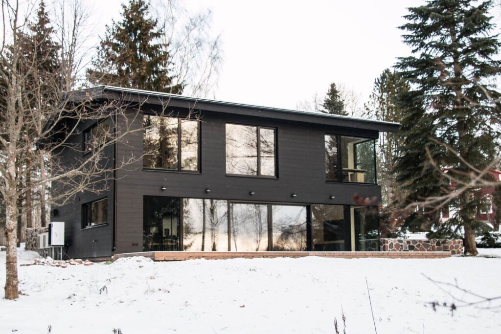 Kahekorruseline musta laudisega modernne puitmaja talvisel maastikul, suurte klaasakendega, ümbritsetud lumiste puude ja kivimüüriga.